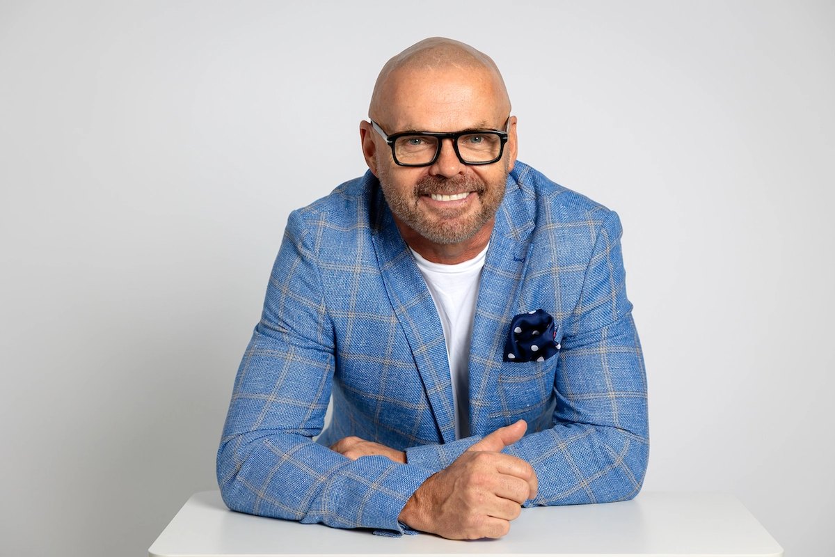 Boss Money founder Tom Uhlich, smiling in a blue checked blazer and glasses, seated at a white desk.