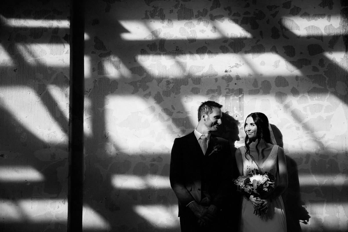 A black-and-white portrait of a bride and groom against a wall dappled with window light.
