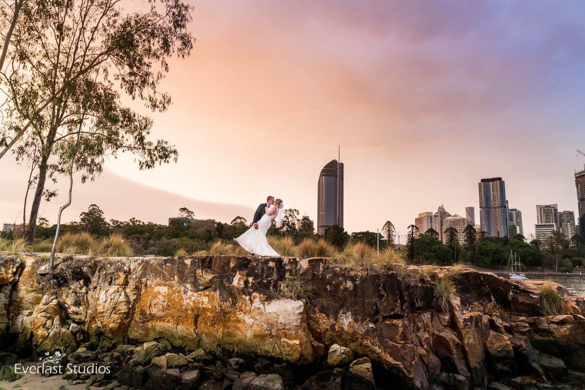 A couple kiss on the Kangaroo Point cliffs at dusk, the Brisbane city skyline rising behind them.