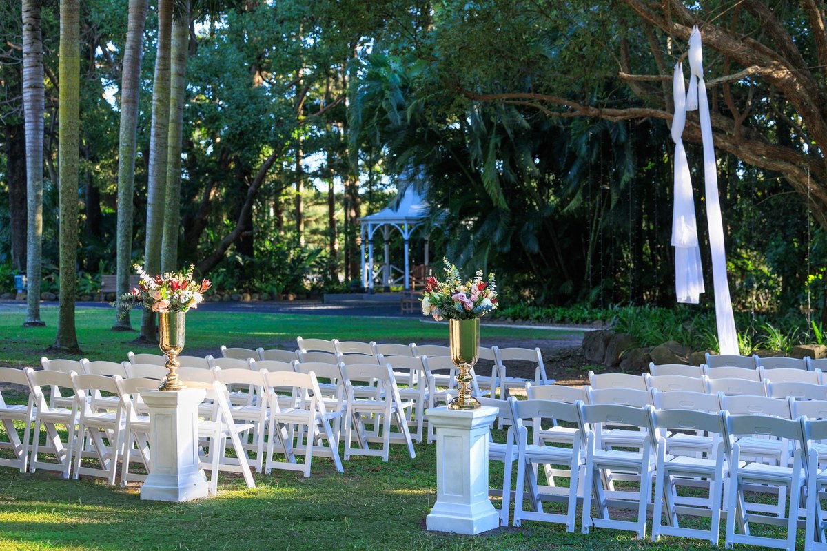 White ceremony chairs and floral pedestals arranged on the lawn, with the Federation gazebo among the trees behind.