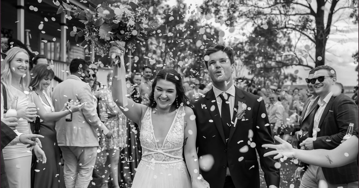A black-and-white photo of a bride and groom walking through a shower of confetti, the bride's bouquet raised high.