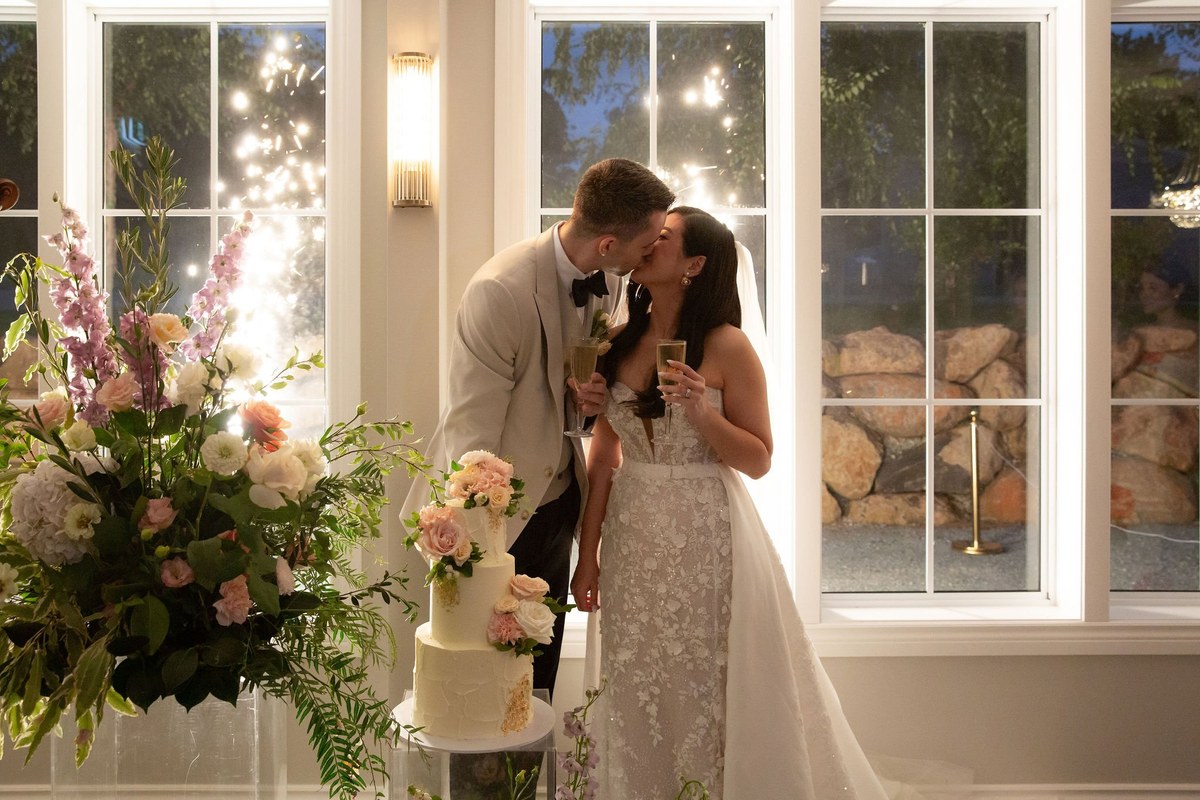 A couple kiss beside their cake in front of the reception hall's French windows as sparklers glow outside.